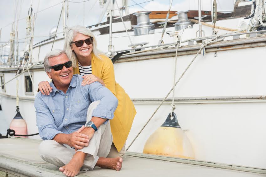 A happy senior couple wearing sunglasses, enjoying a sunny summer day on the waterfront. They are on the boat dock at a marina, smiling, sitting together. They are happily retired or on vacation.