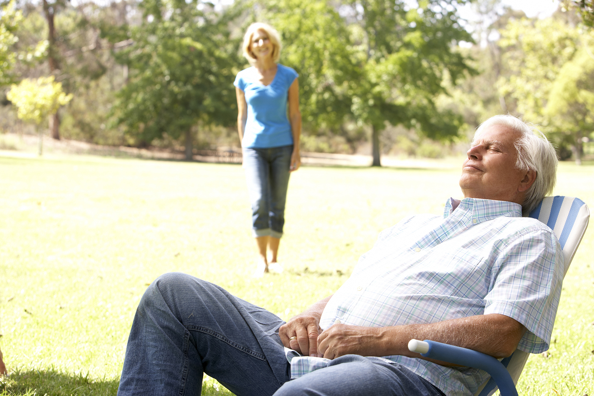 Senior Man Relaxing In Park With Wife In Background